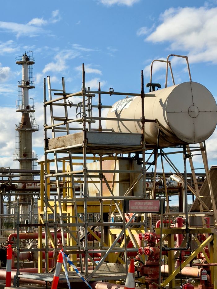 Detailed view of industrial plant infrastructure with scaffolding and storage tank under blue sky.